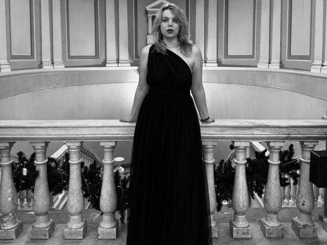 Rosallia stands in black dress, one arm on marble balcony, looking serious. She's got tits, wavy blonde hair, and a sexy, confident vibe. Black-and-white photo.