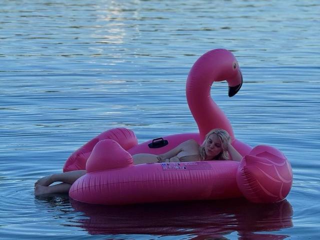 Rosallia floats on a pink inflatable flamingo in a calm, reflective blue water. She's blonde, with fair skin, wearing a black bikini. Her head rests on the flamingo's neck, eyes partially closed. The flamingo's bright pink color contrasts with the water's blue. The inflatable has a textured surface with visible seams. Rosallia's relaxed pose and the smooth water create a serene, leisurely atmosphere. The image uses natural light.