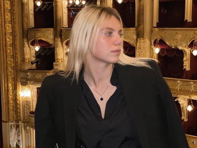 Rosallia with blonde hair, wearing a black blazer and shirt, has a necklace with a small black pendant, looking to the side, in an ornate golden room.