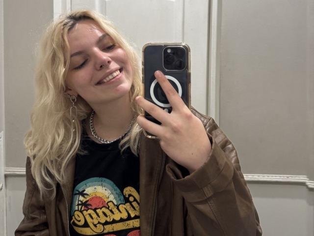 Rosallia with blonde hair, smiling, holding a phone, wearing a brown leather jacket, black shirt, and silver necklace, has a nose ring and hoop earrings, taking a mirror selfie.