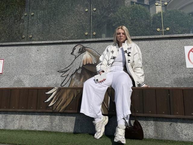 Rosallia sits on a wooden bench against a grey textured wall with a large bird mural. She wears a white bomber jacket with black embroidery, white high-waisted pants, and white sneakers. She has blonde hair and holds a smartphone in her right hand. The mural depicts a detailed bird with spread wings. Rosallia's relaxed posture contrasts with the mural's intricate design. The grassy ground and urban background add depth to the scene.