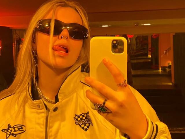 Rosallia with blonde hair, wearing a white sports jacket, black sunglasses, and a silver chain, taking a selfie with her tongue out. Her hand shows a ring and nail polish.