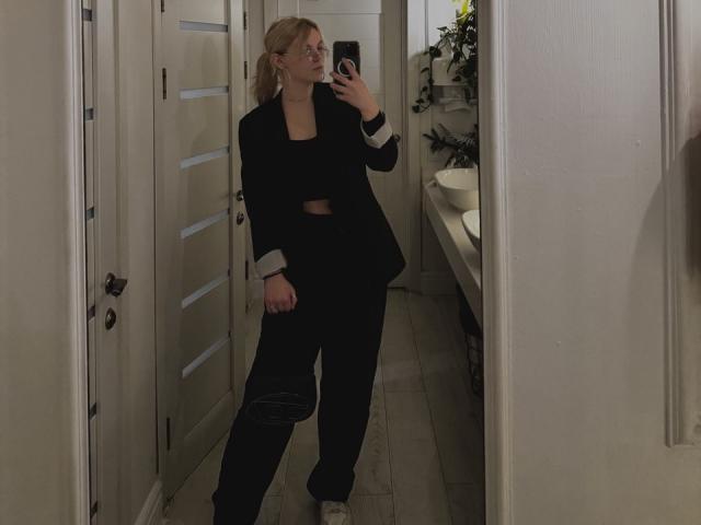 Rosallia stands in a mirror, taking a selfie. She's wearing a black blazer with white cuffs, a black crop top, and black pants. Her blonde hair is tied back, and she holds a black bag in her right hand. The room has a white door with horizontal panels on the left, and a sink with a plant on the right. The floor is light wood. She's looking at her phone with a neutral expression. The image is dimly lit.
