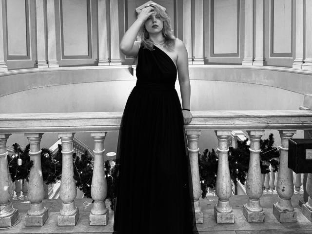 Black and white photo of Rosallia standing on a stone balcony. Rosallia wears a one-shoulder, floor-length black dress, with wavy blonde hair. She's touching her forehead with her right hand. The balcony has ornate, classical-style railings and is adorned with Christmas decorations. Rosallia's dress contrasts with the light, textured background. Her expression is neutral, and the image has a formal, almost cinematic quality.