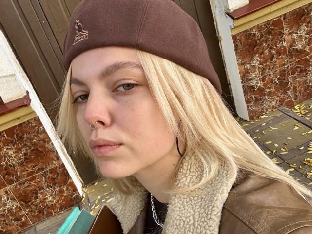 Rosallia with blonde hair, brown beanie, nose ring, and brown leather jacket with shearling collar, standing with a neutral expression, showing a subtle pout.