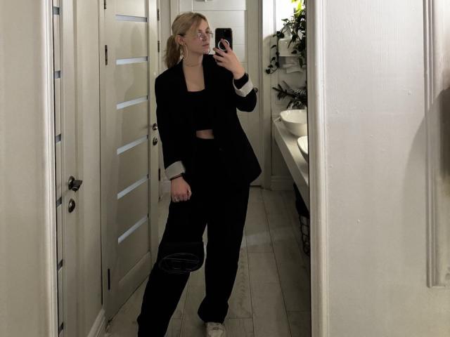 Rosallia stands in a narrow hallway, taking a mirror selfie. She has blonde hair in a ponytail, wears glasses, and a black blazer with white cuffs over a black crop top and black pants. She holds a black bag in her right hand. The floor is light wood, and a bed with white linens is on the right. Rosallia's reflection is clear, and she looks slightly to the left. Her white sneakers are visible. The image is a casual selfie.