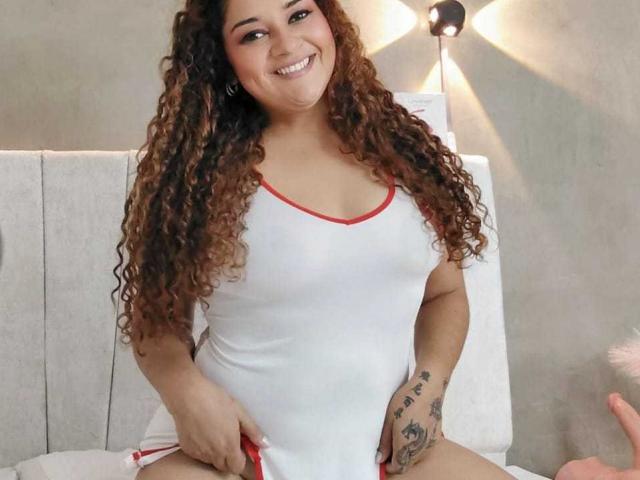 NataliaWendys with curly brown hair, smiling, wearing a white, red-trimmed, tight, see-through dress, showing her tits, and tattoo on right hand, sitting on a bed.