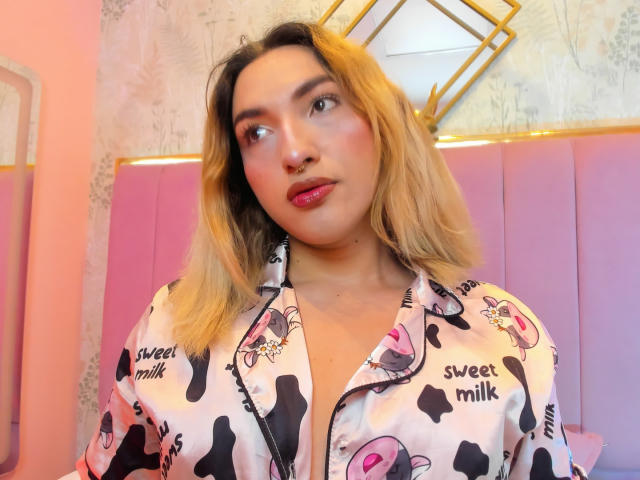TommyTS with blonde hair, pink lips, nose ring, wearing a cow print shirt with "sweet milk" text, looking to the side, pink and purple background.