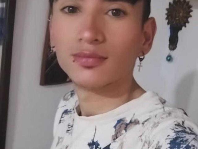 IamSmiith has short dark hair, brown eyes, and a small lip piercing. Wearing a white floral shirt with cross earrings. Soft skin, slight smile, looking at camera.