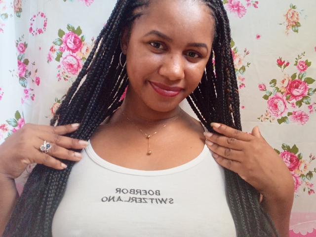 LayahCoquine, a Black woman with long braided hair, smiles at the camera. She wears a white tank top with Russian text, large hoop earrings, and a gold necklace with a small pendant. Her hands, with a ring on the left, hold the tank top's edges. She has a medium build and is set against a floral-patterned background. The image is a photograph.