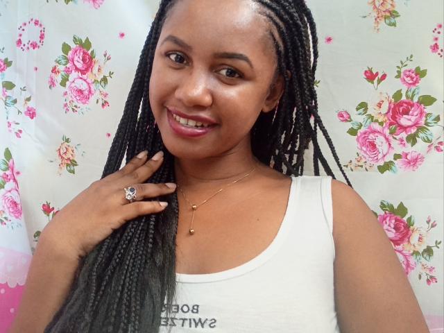 LayahCoquine has dark brown skin and long, straight black braids. She's smiling at the camera with pink lipstick and subtle eye makeup. She's wearing a white tank top and a gold necklace with a small pendant. Her right hand, with a ring on her index finger, touches her braids. The floral pattern in the background is pink and white, with roses and leaves. Her eyebrows are thin and slightly arched. The image is clear and focused on LayahCoquine's upper body.