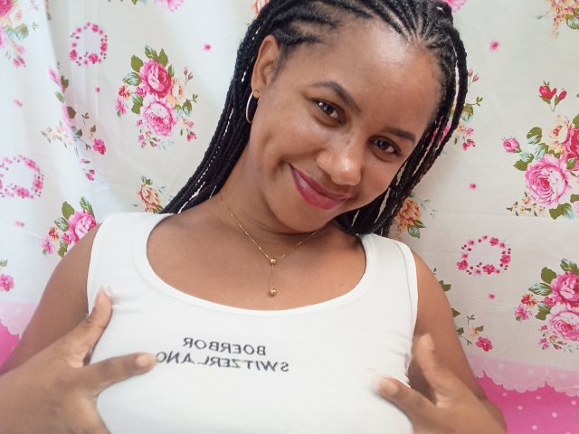 LayahCoquine has dark brown skin and long braided hair. She's smiling at the camera, showing white teeth. She's wearing a white tank top with black text, holding it with both hands. She has small hoop earrings and a gold necklace with a small pendant. Her pink lipstick contrasts with her skin tone. The floral pattern in the background is pink and white. The image has a casual, candid feel.