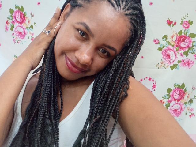 A close-up photo of LayahCoquine, a Black woman with medium-brown skin and long, twisted black braids. She has a slight smile, brown eyes, and is wearing a white tank top. Her right hand touches her hair. She has a gold necklace and small hoop earrings. The background is floral with pink roses on a white pattern. Her braids are neatly arranged, and she looks relaxed. The image is clear and focuses on her face and upper body.