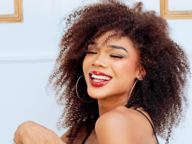 NaomiSteward with curly brown hair, red lipstick, and hoop earrings, smiling with eyes closed, showing white teeth, and wearing a black strap top, looking sexy and happy.