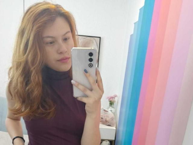 TaylorCarpert with wavy red hair, wearing a maroon turtleneck, taking a selfie in a mirror with a white iPhone, pink and blue striped wall in the background.
