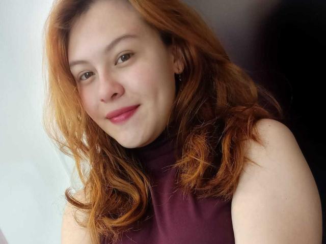 TaylorCarpert has red hair, fair skin, and a slight smile with pink lips. She's wearing a sleeveless maroon top, showing her bare shoulders. Her brown eyes are relaxed.
