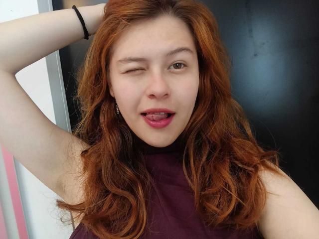 TaylorCarpert, with winking eye and tongue out, has red hair, fair skin, and is wearing a purple sleeveless top, raising her right arm. Black bracelet on her wrist.