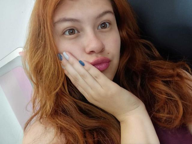 TaylorCarpert with red hair, brown eyes, and pink lips, touching her cheek with blue and white manicured nails, looking at the camera. She's got a small moleskin on her nose.