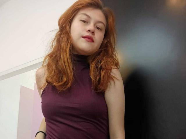 TaylorCarpert has red hair, fair skin, and wears a sleeveless purple top, showing her small tits. She's looking down with a slight smile, pink lips, and a relaxed expression.