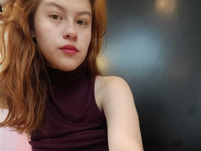 TaylorCarpert with red hair, pale skin, and pink lips, wearing a sleeveless dark purple top, looking slightly bored with a subtle pout. Her bare shoulder is visible.