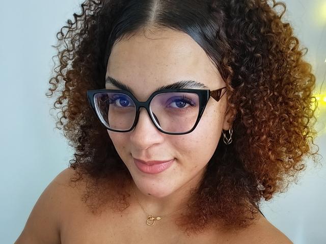 Shaiden with curly brown hair, black glasses, purple eyeshadow, and a subtle smile, showing off her small, perky tits, and a gold necklace against a plain background.