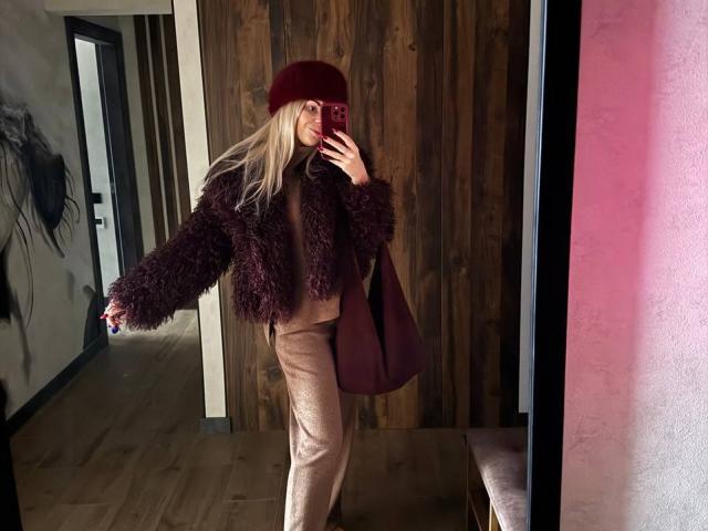 BellaRayyy with long blonde hair, wearing a dark purple furry jacket, red fur hat, beige pants, holding a red phone, standing in a wooden-walled room, showing her ass.