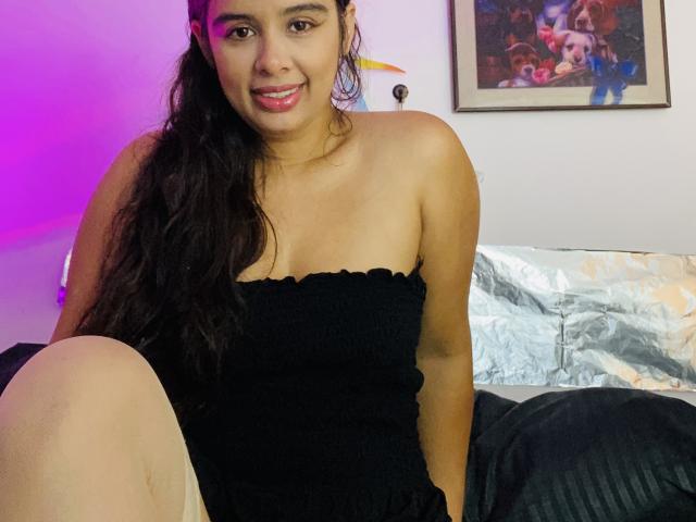 KeyklinOne tiene la piel morena, el pelo largo y negro, y está sonriendo con los labios rosados. Lleva un top negro sin tirantes, está sentada con una rodilla levantada, mostrando su trasero desnudo..
