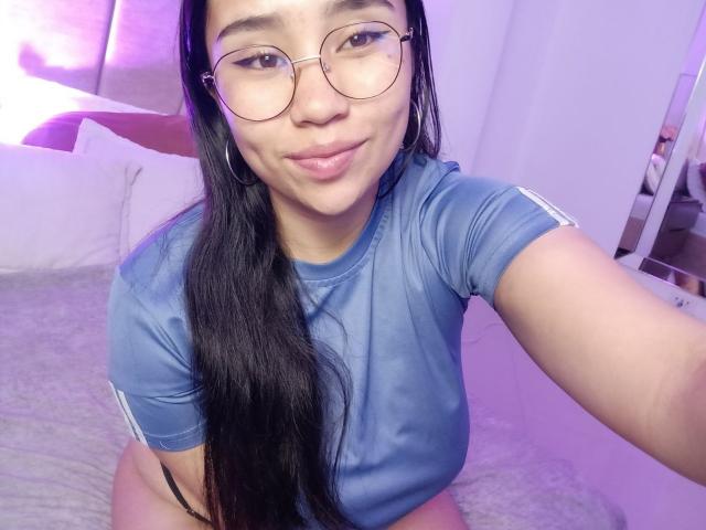 Karollteyes with long black hair, glasses, and hoop earrings, wearing a blue shirt, sitting on a bed, showing a slight smile, and exposing her bare ass.