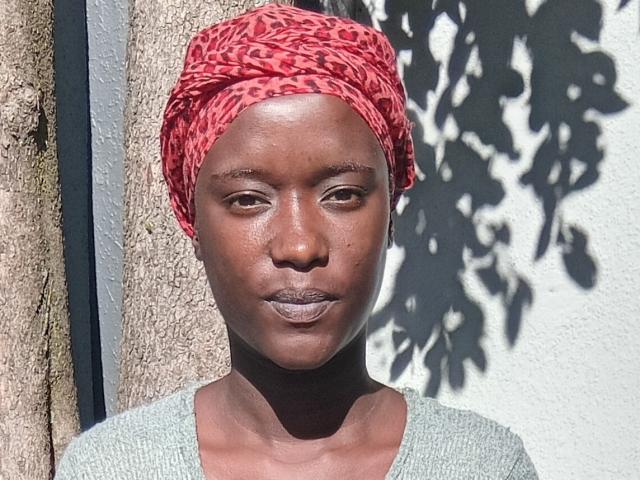 BlackFirre with dark brown skin, wearing a red leopard-print headscarf, stands in front of a textured wall. She has a neutral expression, small nose, and slightly parted lips. She wears a light gray, knit top. Sunlight casts a shadow of tree leaves on her face and headscarf. Her head is slightly tilted to the right. The texture of the wall and headscarf contrast with her smooth skin. Image has a realistic, natural style.