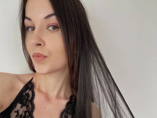JuliaSexxx-hot has long black hair, light skin, pink eyeshadow, and is wearing a black lace halter top, with a neutral expression. Her tits are partially visible.