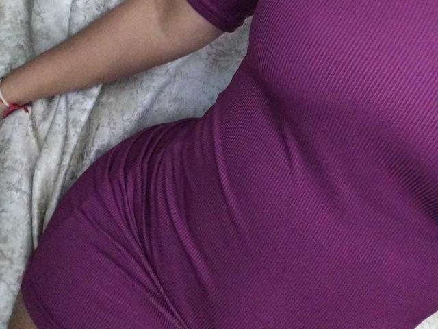 AriaVega's purple ribbed dress hugs her curvy ass and small boobs, with her right arm resting on a patterned surface. She wears a red bracelet.