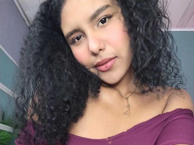 AriaVega with curly black hair, light brown skin, small nose, and pouty pink lips, wearing an off-shoulder purple top, necklace, and subtle makeup, looking at camera.