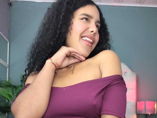 AriaVega with curly black hair, light brown skin, smiling, and looking to the side. She wears an off-shoulder purple top, revealing a bit of her collarbone. Her right hand rests on her chin, showing a small red bracelet. She has a delicate necklace. The background is a teal wall with a potted plant and a pink-lit corner. The composition is slightly tilted.