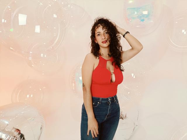 AmaraYasbel has curly brown hair, red sleeveless top with keyhole cutout showing her boobs, and blue high-waisted jeans, standing with one hand in her hair, smiling.