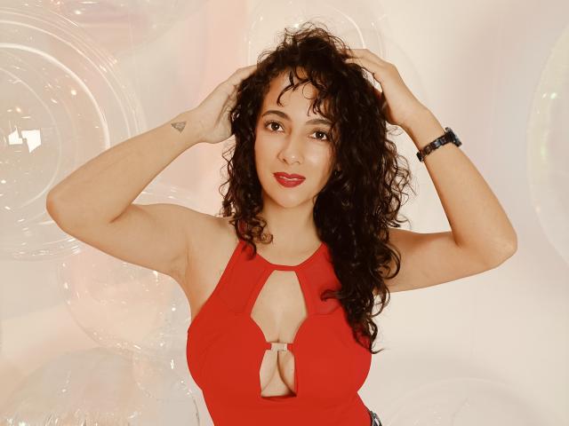 AmaraYasbel has curly black hair, red lipstick, and wears a red cutout top showing her tits. She's smiling, hands in her hair, with a small triangle tattoo.