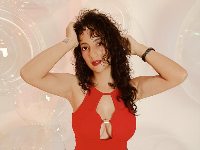 AmaraYasbel with curly dark hair, red cutout top showing small tits, hands in hair, red lipstick, tattoo on right wrist, bracelet on left wrist, smiling, standing.