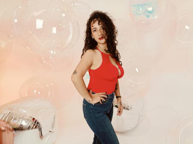 AmaraYasbel with curly brown hair, red tank top, and blue jeans, showing off her big boobs and tattoo on her right arm, standing confidently with hand on hip.