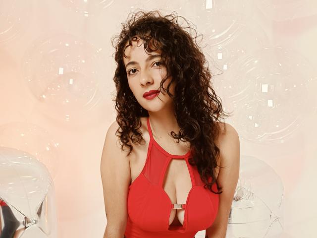 AmaraYasbel with curly brown hair, red cleavage dress, showing her tits, sitting in front of transparent balloons, red lipstick, slightly smiling, soft pink background.