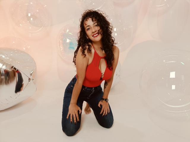 AmaraYasbel kneeling, curly brown hair, red halter top with cutout, showing her tits, dark blue jeans, smiling, hands on knees, silver balloon on left, white background.
