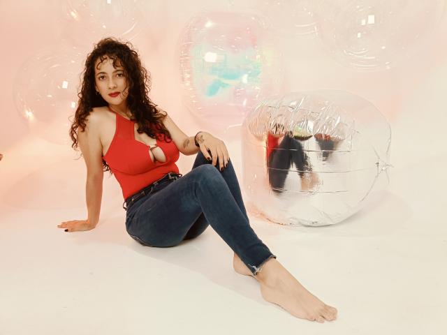 AmaraYasbel with curly brown hair, red cleavage top, blue jeans, barefoot, sitting, legs crossed, surrounded by transparent balloons and a reflective silver ball, smiling.