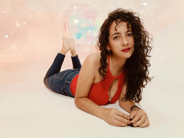 AmaraYasbel, curly-haired, laying on her stomach, red top, showing small tits, denim jeans, bare feet, red lipstick, white background, bubbles in background, relaxed, slightly smiling.