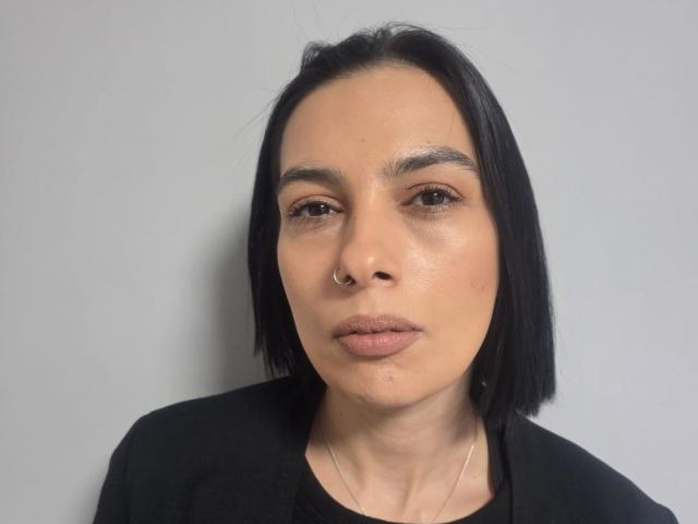 LillyyMoon has a nose ring, black straight bob, and a neutral expression with slightly parted lips. She's wearing a black top and a thin necklace.