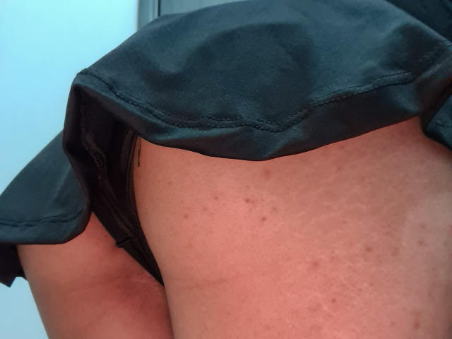 NaylaGrey's black leather skirt is lifted, exposing her bare ass and black panties. Her skin has small freckles and a few blemishes. Her right thigh is visible.