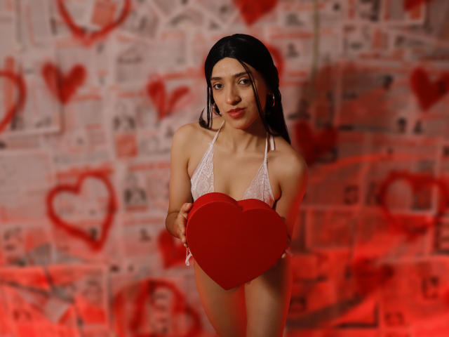 MichaelaMistress with long black hair, wearing a white lace bikini, holding a red heart, standing in a red-lit room, looking at the camera with a slight smile.