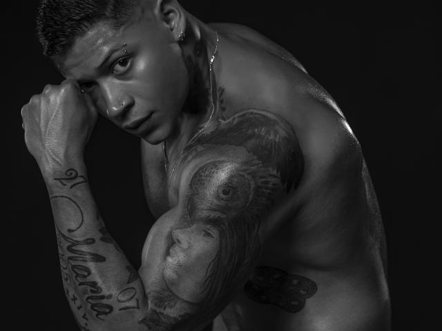 DiagoRockfield has a muscular, tattooed upper body with a detailed bird tattoo on his right arm, short hair, and a nose ring, looking serious in a black-and-white photo.