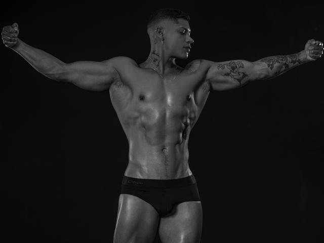 DiagoRockfield, muscular, shirtless, black briefs, arms spread, tattoos on arm and neck, short hair, glistening skin, standing, dark background, grey-scale, defined abs, visible bulge, serious expression.