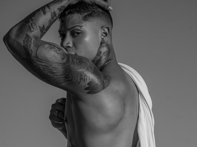 DiagoRockfield with tattoos, muscular arms, and a short haircut, wearing a draped white cloth, hand on head, looking back, gray-scale photo. DiagoRockfield's butt is visible from the side.