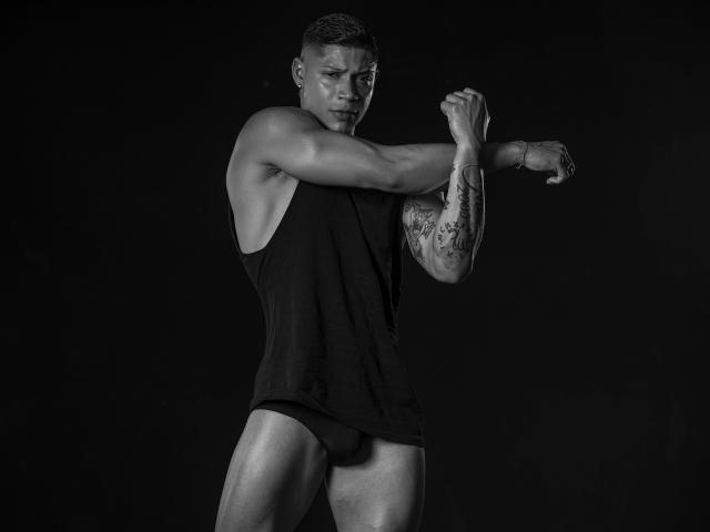 DiagoRockfield, muscular black dude, sleeve tattoo, short buzzcut, black tank top, black underwear, stretching, intense gaze, strong arms, visible bulge, black and gray photograph.