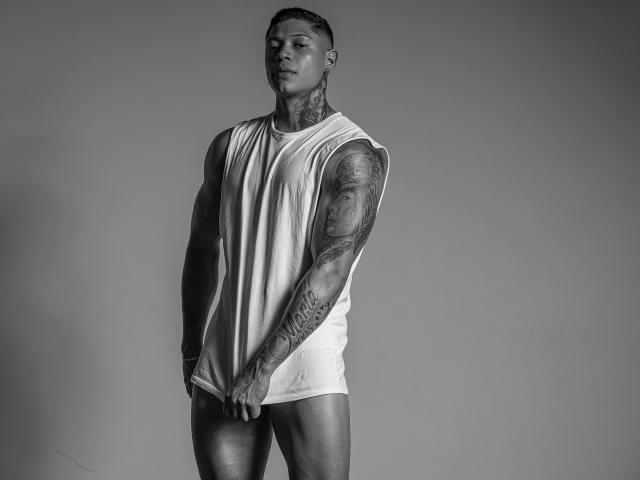 DiagoRockfield, a muscular tattooed dude with short hair, wearing a sleeveless white tank top, showing his butt, standing with a serious expression, holding his tank top. Black-and-white photo.
