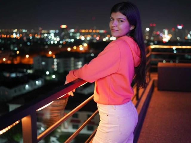RilleyFoyx with long dark hair, pink hoodie, and white pants, standing on a balcony at night, looking over her shoulder, holding a railing, city lights in the background.